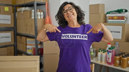 Photo for Middle age hispanic woman pointing to volunteer uniform smiling at charity center - Royalty Free Image