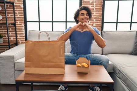 Photo for Hispanic man with curly hair eating chicken wings smiling in love doing heart symbol shape with hands. romantic concept. - Royalty Free Image