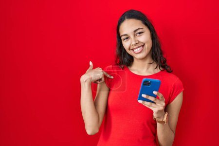 Foto de Mujer brasileña joven usando teléfono inteligente sobre fondo rojo mirando confiado con sonrisa en la cara, señalándose con los dedos orgullosos y felices. - Imagen libre de derechos