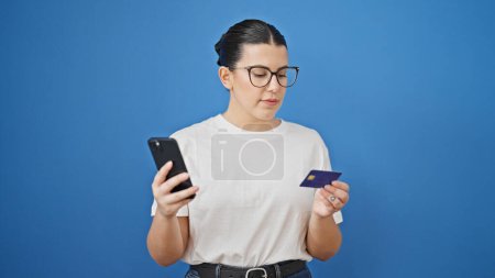 Téléchargez les photos : Jeune belle femme hispanique utilisant smartphone et carte de crédit sur fond bleu isolé - en image libre de droit