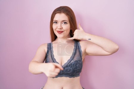 Photo for Redhead woman wearing lingerie over pink background smiling doing talking on the telephone gesture and pointing to you. call me. - Royalty Free Image