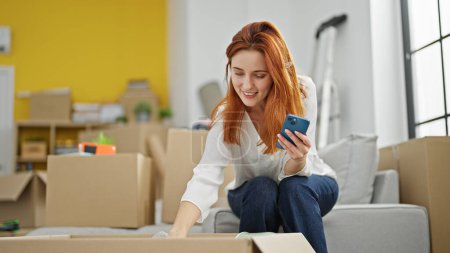 Foto de Mujer pelirroja joven desempacando la caja de cartón utilizando el teléfono inteligente en el nuevo hogar - Imagen libre de derechos
