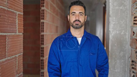 Téléchargez les photos : Jeune homme hispanique travailleur debout avec un visage sérieux sur le chantier de construction - en image libre de droit