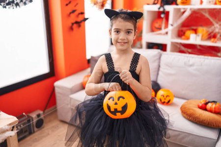Téléchargez les photos : Adorable fille hispanique portant costume d'Halloween tenant panier de citrouille à la maison - en image libre de droit