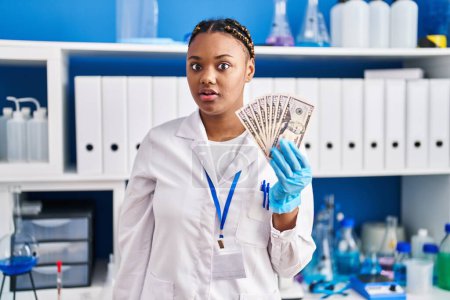Foto de Mujer afroamericana con trenzas trabajando en laboratorio científico sosteniendo dinero asustada y sorprendida con la boca abierta para sorpresa, cara de incredulidad - Imagen libre de derechos