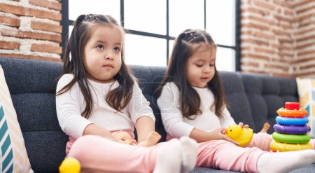 Téléchargez les photos : Adorable jumelles jouant avec cerceaux jeu tenant jouet de canard assis sur le canapé à la maison - en image libre de droit