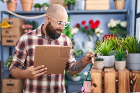 Téléchargez les photos : Young bald man florist reading document holding plant at flower shop - en image libre de droit