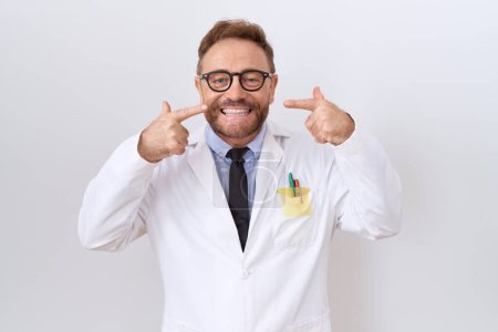 Téléchargez les photos : Médecin d'âge moyen homme avec barbe vêtu d'un manteau blanc souriant gai montrant et pointant avec les doigts des dents et de la bouche. concept de santé dentaire. - en image libre de droit