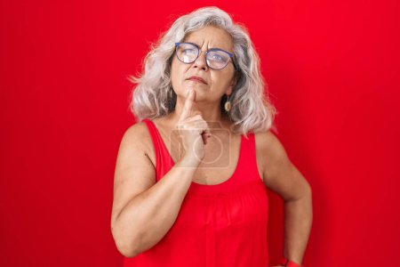 Téléchargez les photos : Femme d'âge moyen avec les cheveux gris debout sur fond rouge pensant concentré sur le doute avec le doigt sur le menton et regardant vers le haut se demandant - en image libre de droit