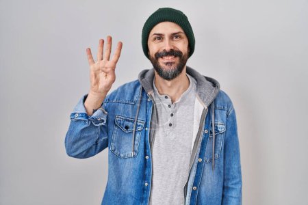 Foto de Joven hombre hispano con tatuajes con gorra de lana mostrando y señalando hacia arriba con los dedos número cuatro mientras sonríe confiado y feliz. - Imagen libre de derechos