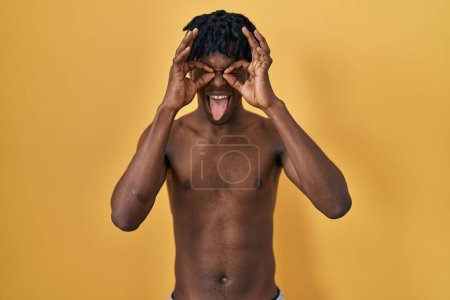 Téléchargez les photos : Jeune homme africain avec dreadlocks debout torse nu faisant un geste correct comme des jumelles sortant la langue, les yeux regardant à travers les doigts. expression folle. - en image libre de droit