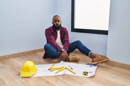Photo for African american man sitting on the floor at new home looking at blueprints puffing cheeks with funny face. mouth inflated with air, crazy expression. - Royalty Free Image