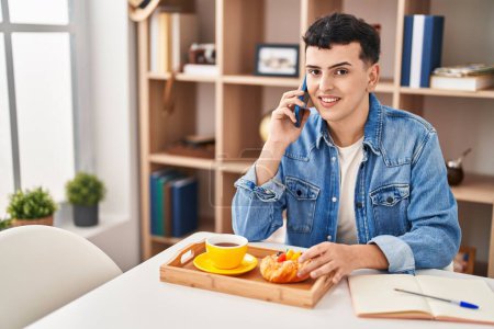 Téléchargez les photos : Jeune homme non binaire parlant sur le smartphone prendre le petit déjeuner à la maison - en image libre de droit