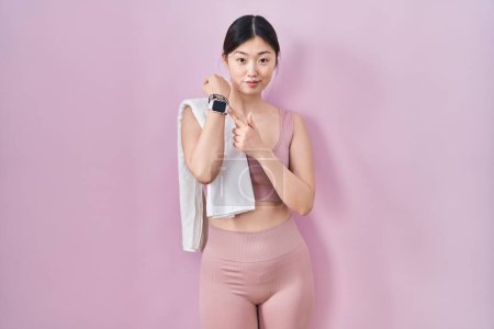 Téléchargez les photos : Jeune femme chinoise portant des vêtements de sport et une serviette en hâte pointant vers regarder le temps, l'impatience, regarder la caméra avec une expression détendue - en image libre de droit