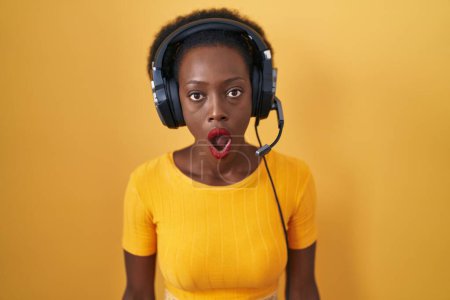 Photo for African woman with curly hair standing over yellow background wearing headphones afraid and shocked with surprise expression, fear and excited face. - Royalty Free Image