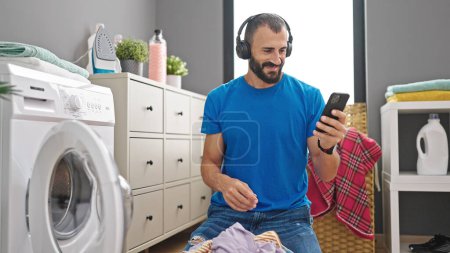 Téléchargez les photos : Jeune homme hispanique laver les vêtements à l'aide d'un smartphone et d'écouteurs à la buanderie - en image libre de droit