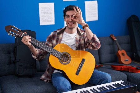 Foto de Joven hombre hispano tocando la guitarra clásica en el estudio de música sonriendo feliz haciendo signo bien con la mano en el ojo mirando a través de los dedos - Imagen libre de derechos