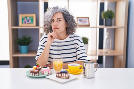 Foto de Mujer de mediana edad con cabello gris comiendo pasteles y tomando café para el desayuno cara seria pensando en la pregunta con la mano en la barbilla, reflexivo acerca de la idea confusa - Imagen libre de derechos