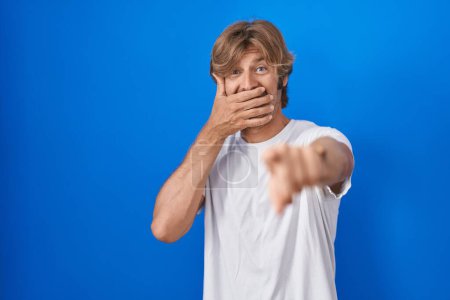 Photo for Middle age man standing over blue background laughing at you, pointing finger to the camera with hand over mouth, shame expression - Royalty Free Image