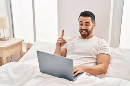 Foto de Joven hispano con barba usando computadora portátil en la cama sonriendo con una idea o pregunta apuntando con el dedo con la cara feliz, número uno - Imagen libre de derechos