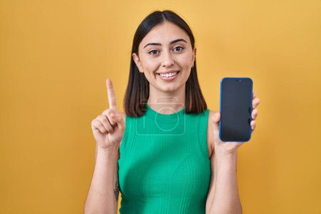 Foto de Chica hispana sosteniendo teléfono inteligente mostrando la pantalla sonriendo con una idea o pregunta señalando el dedo con la cara feliz, número uno - Imagen libre de derechos