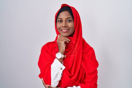 Téléchargez les photos : Jeune femme arabe portant l'écharpe traditionnelle islamique hijab avec la main sur le menton penser à la question, expression coûteuse. Visage souriant et réfléchi. concept de doute. - en image libre de droit