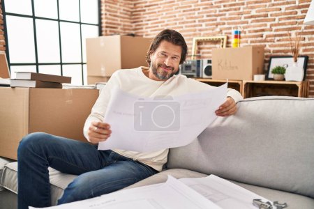 Téléchargez les photos : Homme d'âge moyen souriant confiant regardant des plans de maison à la nouvelle maison - en image libre de droit