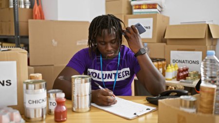 Téléchargez les photos : Un homme afro-américain bénévole écoutant un message vocal écrire sur un document au centre caritatif - en image libre de droit