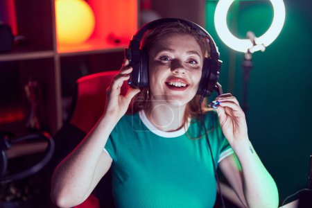 Téléchargez les photos : Jeune rousse femme streamer souriant confiant assis sur la table à la salle de jeux - en image libre de droit