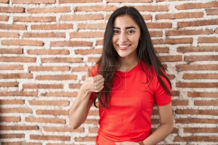 Téléchargez les photos : Jeune adolescente debout sur un mur de briques faisant des pouces heureux geste avec la main. approuvant l'expression regardant la caméra montrant le succès. - en image libre de droit