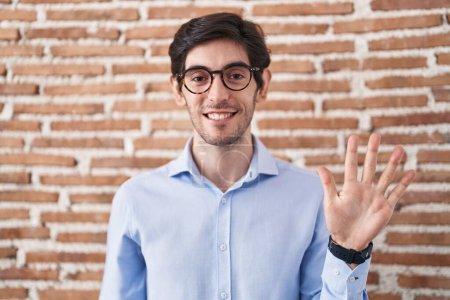 Téléchargez les photos : Jeune homme hispanique debout sur fond de mur de briques montrant et pointant vers le haut avec les doigts numéro cinq tout en souriant confiant et heureux. - en image libre de droit