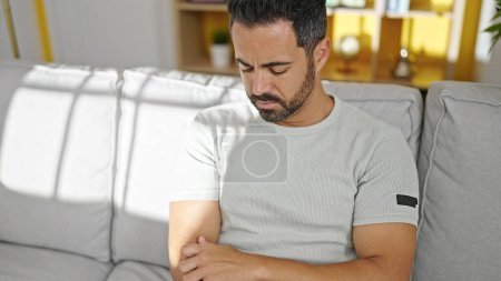 Téléchargez les photos : Jeune homme hispanique assis sur le canapé bras à gratter à la maison - en image libre de droit