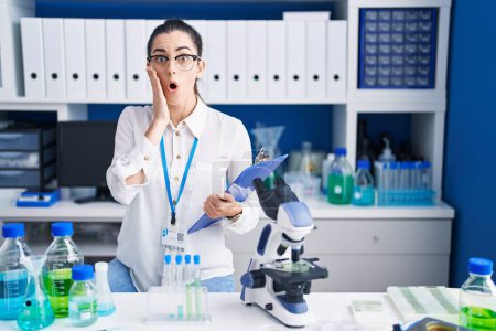 Téléchargez les photos : Jeune femme brune travaillant au laboratoire scientifique effrayée et choquée, surprise et expression étonnante avec les mains sur le visage - en image libre de droit