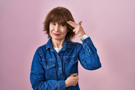 Photo for Middle age woman standing over pink background shooting and killing oneself pointing hand and fingers to head like gun, suicide gesture. - Royalty Free Image