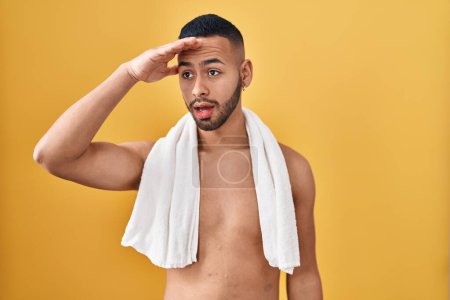 Photo for Young hispanic man standing shirtless with towel very happy and smiling looking far away with hand over head. searching concept. - Royalty Free Image