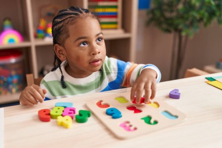 Téléchargez les photos : Africain américain garçon jouer avec des mathématiques jeu de puzzle assis sur la table à la maternelle - en image libre de droit
