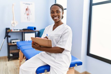 Photo for African american woman physiotherapist smiling confident writing medical report at rehab clinic - Royalty Free Image