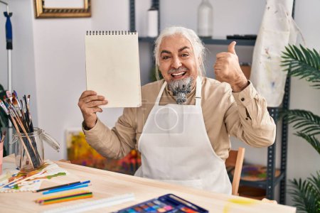 Téléchargez les photos : Homme d'âge moyen aux cheveux gris assis au studio d'art tenant un cahier pointant le pouce vers le côté souriant heureux avec la bouche ouverte - en image libre de droit
