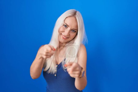 Foto de Mujer caucásica de pie sobre fondo azul señalando los dedos a la cámara con la cara feliz y divertida. buena energía y vibraciones. - Imagen libre de derechos