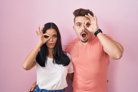 Foto de Joven pareja hispana de pie sobre fondo rosa haciendo un gesto bien sorprendido con la cara sorprendida, los ojos mirando a través de los dedos. expresión incrédula. - Imagen libre de derechos