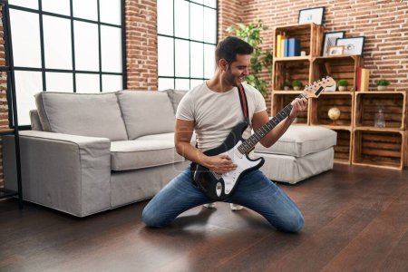Photo for Young hispanic man playing electrical guitar with knees on floor at home - Royalty Free Image