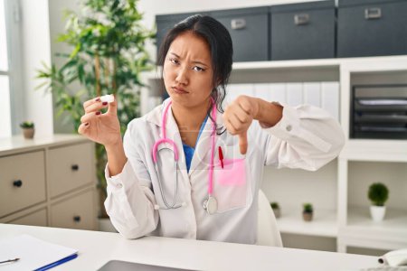 Photo for Young asian woman wearing doctor uniform and stethoscope with angry face, negative sign showing dislike with thumbs down, rejection concept - Royalty Free Image