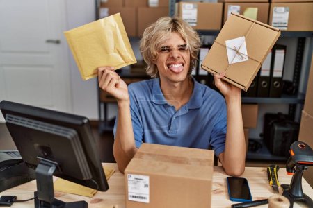 Téléchargez les photos : Young man working at small business ecommerce holding packages sticking tongue out happy with funny expression. - en image libre de droit
