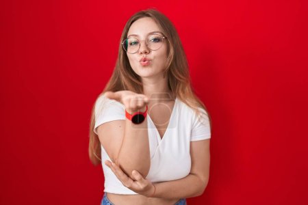 Photo for Young caucasian woman standing over red background looking at the camera blowing a kiss with hand on air being lovely and sexy. love expression. - Royalty Free Image