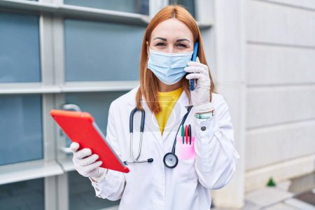 Téléchargez les photos : Jeune femme caucasienne médecin portant un masque médical en utilisant touchpad parler sur smartphone à l'hôpital - en image libre de droit