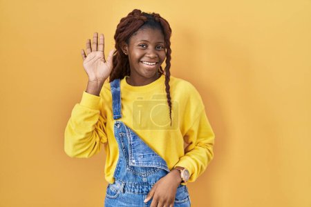 Téléchargez les photos : Femme africaine debout sur fond jaune renonçant à dire bonjour heureux et souriant, geste de bienvenue amical - en image libre de droit