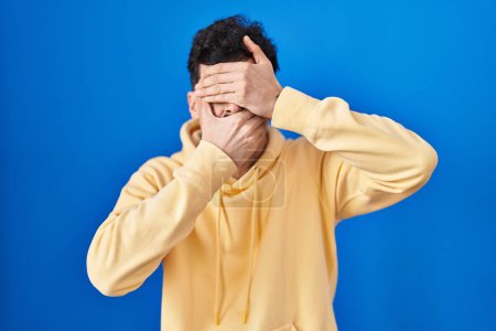 Photo for Hispanic man standing over blue background covering eyes and mouth with hands, surprised and shocked. hiding emotion - Royalty Free Image