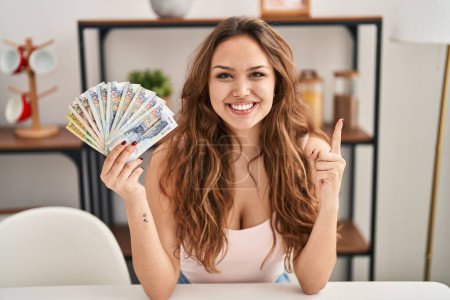 Photo for Young hispanic woman holding peruvian sol banknotes smiling with an idea or question pointing finger with happy face, number one - Royalty Free Image