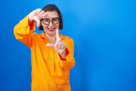 Téléchargez les photos : Femme hispanique du Moyen Âge portant des lunettes debout sur fond bleu souriant faisant cadre avec les mains et les doigts avec le visage heureux. concept de créativité et photographie. - en image libre de droit