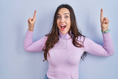 Photo for Young brunette woman standing over blue background smiling amazed and surprised and pointing up with fingers and raised arms. - Royalty Free Image